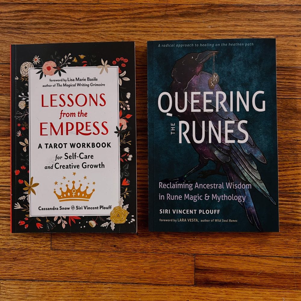 Two books next to each other on a hardwood floor. On the left is "Lessons from the Empress: a tarot workbook for self-care and creative growth." It has a simple cover with a floral design in different shades of orange on a black background. "Queering the Runes" is on the right, and the cover is a textured dark blue background with a raven with slight, dark rainbow feathers. The raven is holding two runes on strings in its beak: Dagaz and Ingwaz.