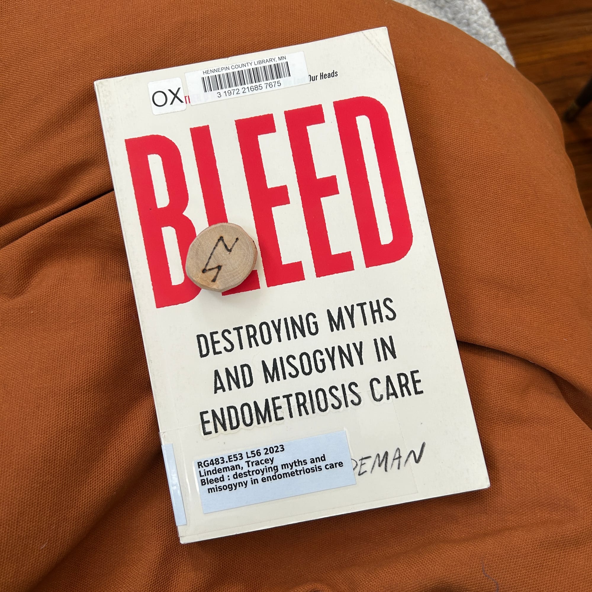 A copy of "Bleed: Destroying Myths and Misogyny in Endometriosis Care" on top of an orange pillow. The Perthro rune rests on top of it.